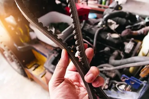 4 Timing Belt Warning Signs To Avert Disaster | EG Auto Center in Dayton, NJ. Image of a mechanic’s hand holding a timing belt with a blurred car engine in the background.